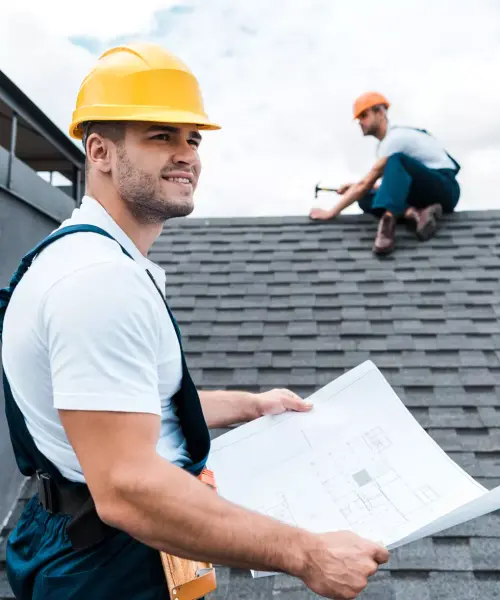 Two roofers working on a house roof, one holding blueprints.